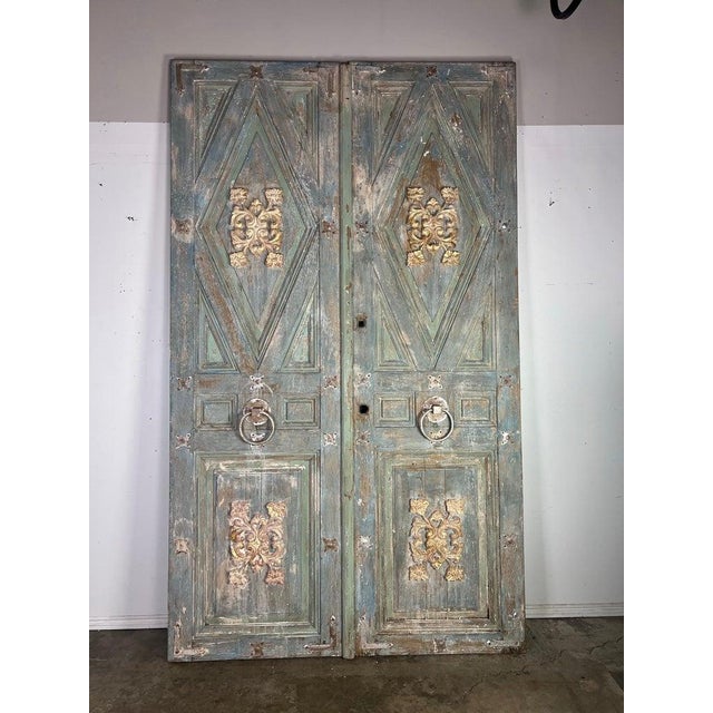 Pair of tall, rustic double doors with an ornate and distressed finish. The doors are painted in a faded greenish-blue...