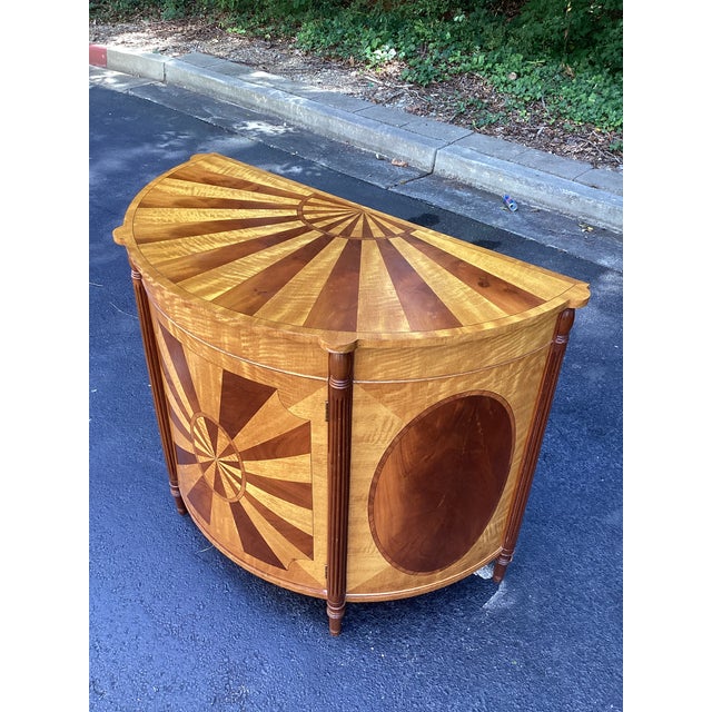 Striking Neoclassical-style demilune cabinet featuring elaborate sunburst marquetry in contrasting warm-toned woods. The...
