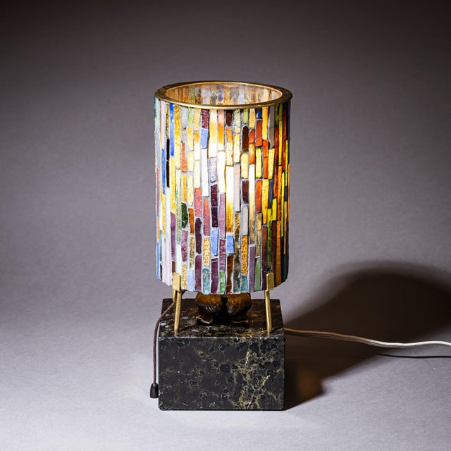 Small table lamp on a black marble base with brass fittings and a cylindrical glass shade composed of differently colored...