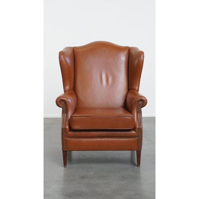 Offered is this stylish and in correct condition cognac colored cowhide leather English style ear chair with decorative...