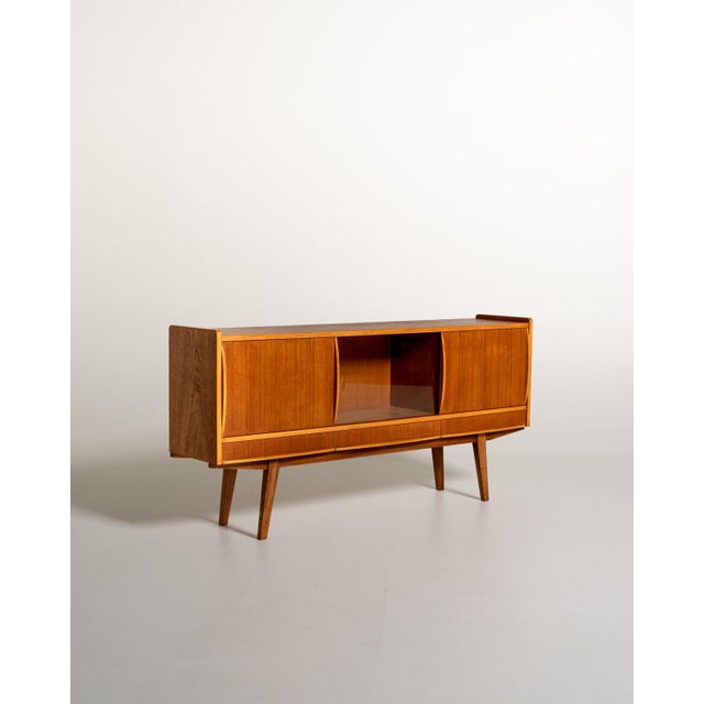 Teak frame with shaped sliding doors, a central compartment with smoked glass, three lower drawers, and slanted dark wood...
