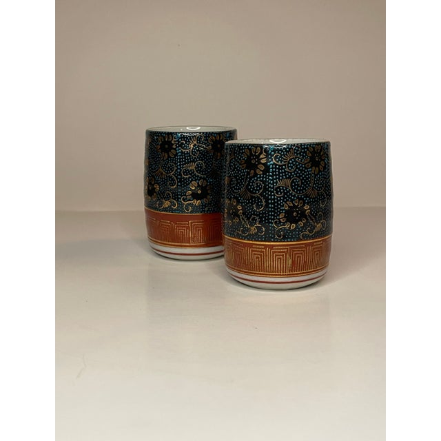 A pair of white ceramic glasses with a pattern of black & gold floral scrollwork on a rich green speckled background...