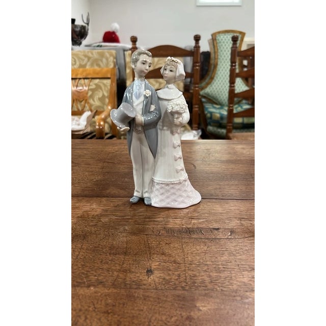 An elegant vintage porcelain figurine depicting a newlywed couple in classic Lladró-inspired soft pastel tones. The groom...