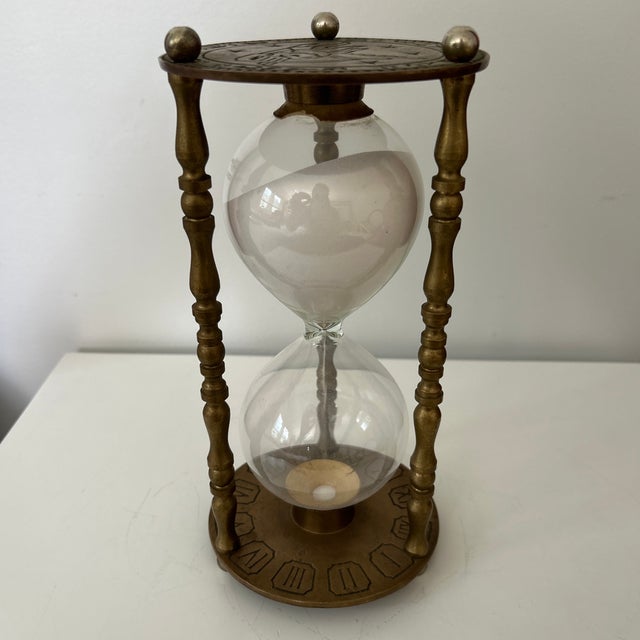 A distinctive vintage hourglass featuring a clear glass timer filled with white sand and held within an ornate three-...