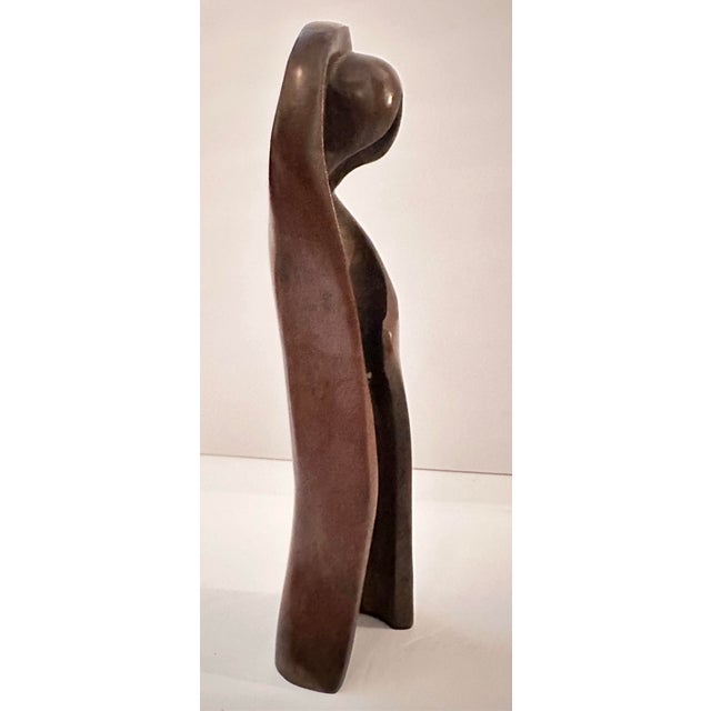 A tall, abstracted bronze figure, built from one continuous sweeping curve that loops up and around to form a rounded head...