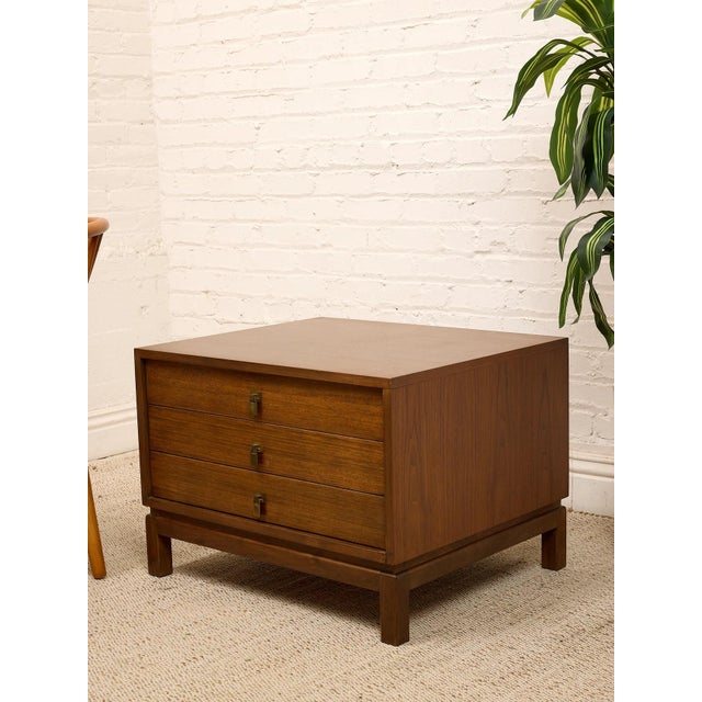 A handsome vintage walnut nightstand featuring three spacious drawers, warm wood grain, and distinctive brass campaign-...
