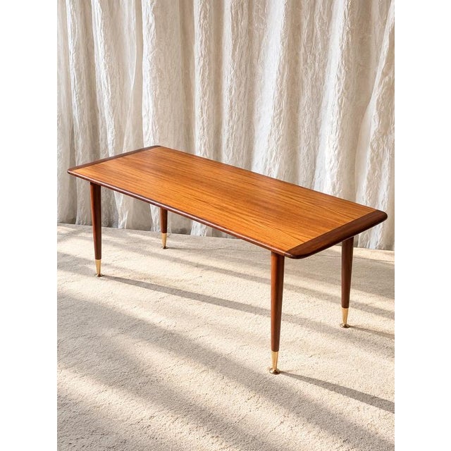 Elegant Mid-Century Modern coffee table, circa 1960, combining warm-toned teak with refined brass details. The elongated...