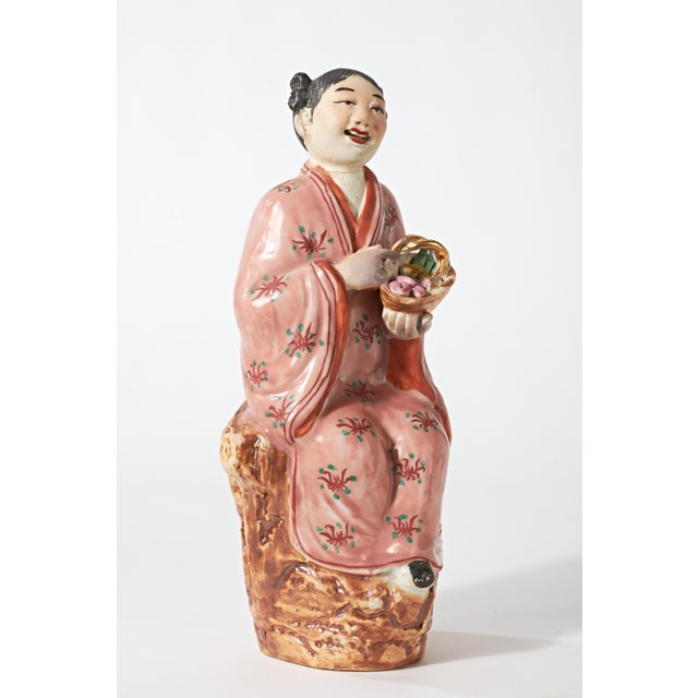 Small hand painted porcelain figurine of young Chinese woman in brightly colored floral pink attire. She is seated on a...