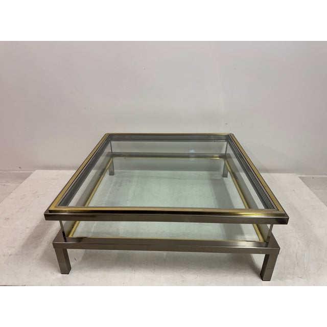 A low, square vitrine coffee table in polished metal with a restrained, architectural profile. The frame is a combination...