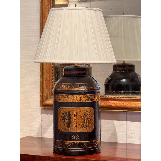 Tea tin lamp in black paint with gilt bands and a gilt panel of characters, numbered 32, fitted with a pleated shade.