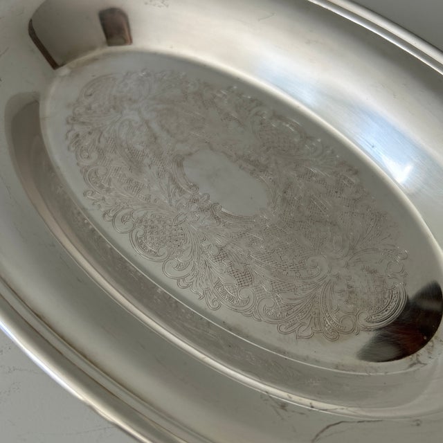 Elegant vintage silver-plated oval serving platter featuring an ornate engraved floral and scrollwork medallion at the...