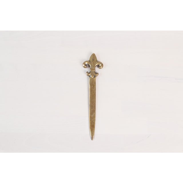 A vintage brass letter opener with a cast fleur-de-lys handle and tapered blade. The brass has a warm aged patina with...