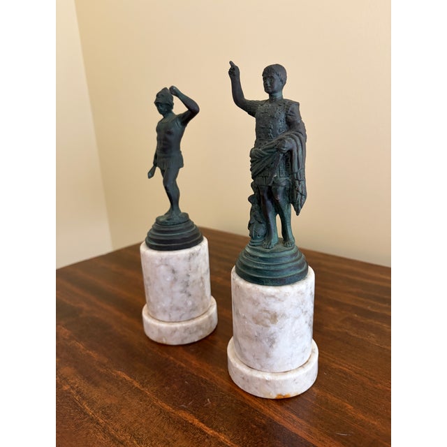 A pair of Grand Tour–style classical figures in brass, mounted on a cylindrical marble bases. The slightly more relaxed...