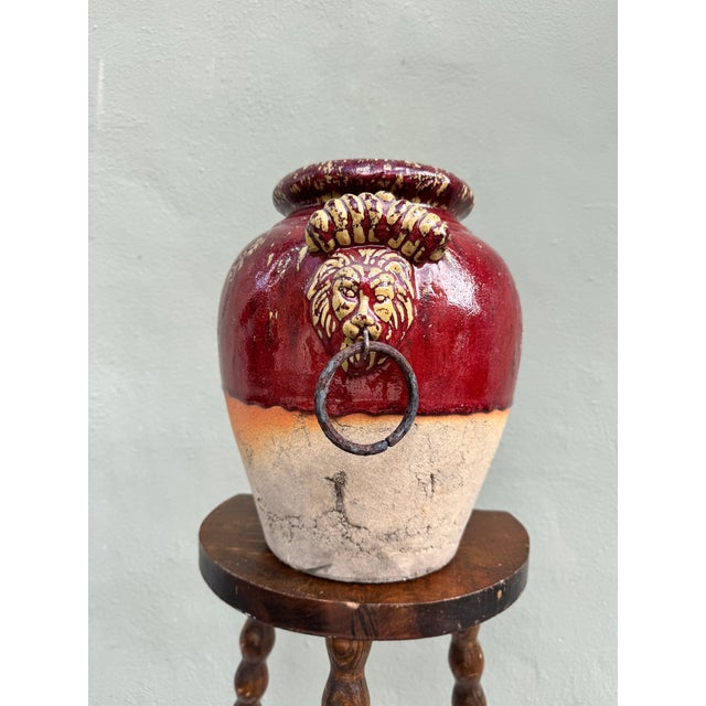 A commanding terracotta vessel with sculptural lion-head handles, finished in rich red and cream crackle glaze. Evokes...