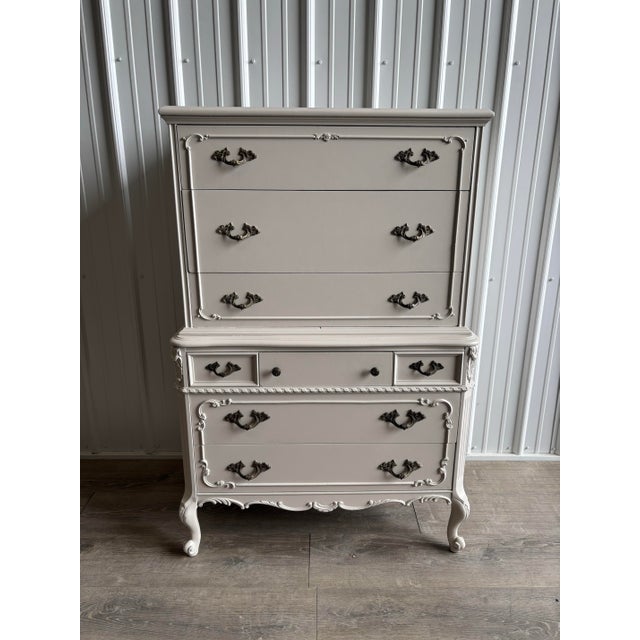 An elegant French Provincial tallboy dresser featuring graceful curves, ornate carved detailing, and a soft neutral finish...