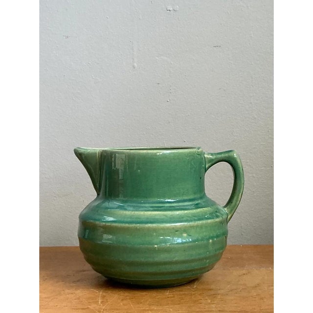 This vintage green glazed ceramic pitcher presents a charming, squat silhouette, with a full, rounded belly that rises to...