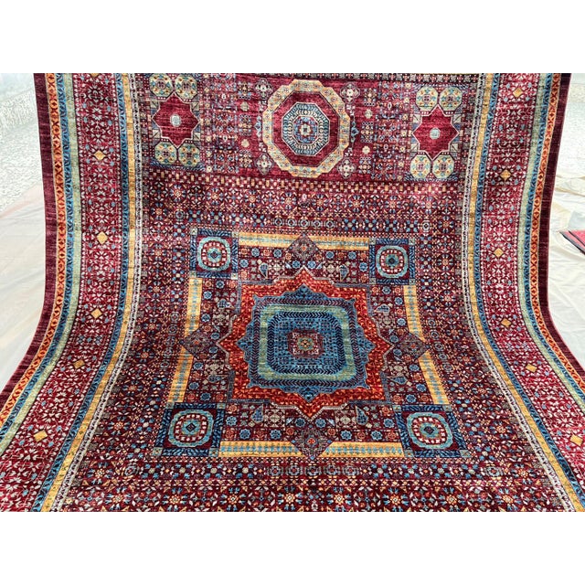 This Mamluk-style wool area rug features a rich red field with geometric medallions, detailed floral motifs, and...