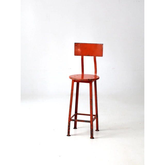 This is a vintage industrial stool. The orange metal stool features a round wooden seat and an arched back rest.