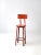 This is a vintage industrial stool. The orange metal stool features a round wooden seat and an arched back rest.