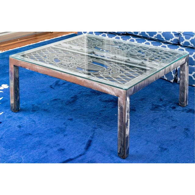 Vintage well-crafted glass top coffee table with a heavy brush steel based with an open fretwork of dual panels of floral...