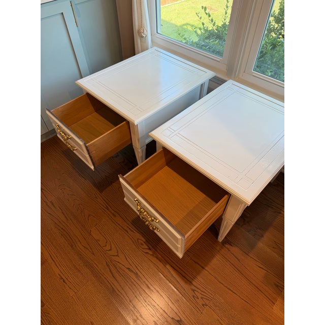 An elegant matching pair of Brandt nightstands featuring a white painted finish, decorative gold-tone drawer pulls, and...