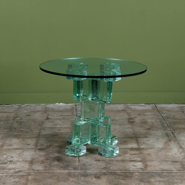 .Postmodern occasional table is anchored by a heavy, geometric pedestal base composed of interlocking, solid cast-glass...