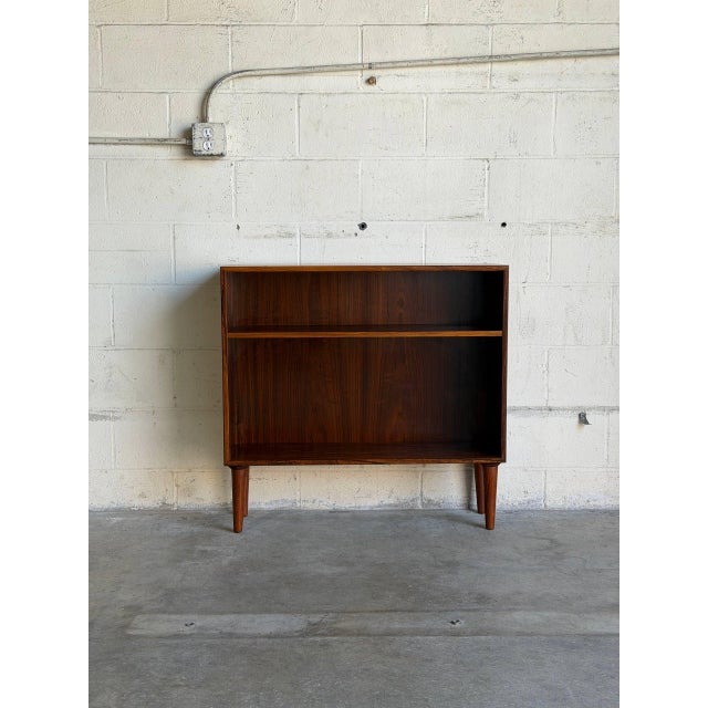This compact rosewood bookcase pairs a clean rectangular case with one open shelf and two display levels. Four tapered...