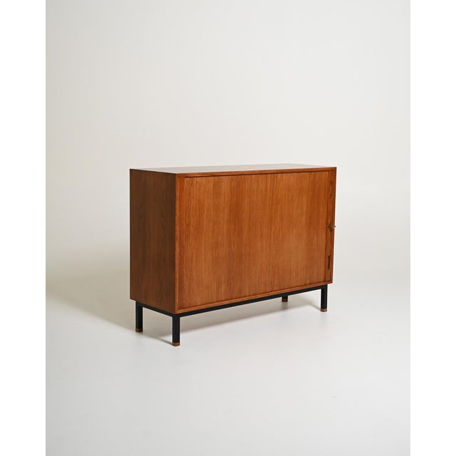 Oak cabinet with sliding roller door, adjustable shelves, and black lacquered base. Scandinavian design, 1960s. The...