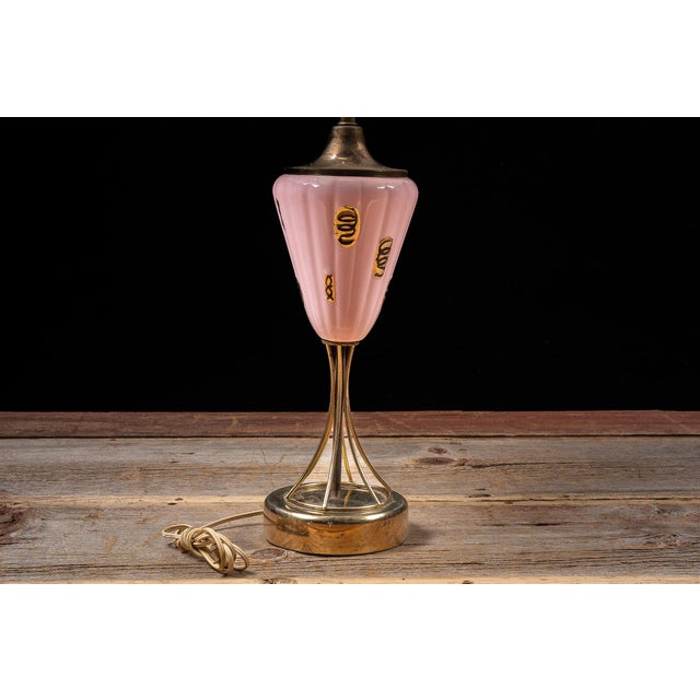 Mid-century American table lamp featuring a tapered ribbed pink glass body decorated with gilt motifs and supported by an...