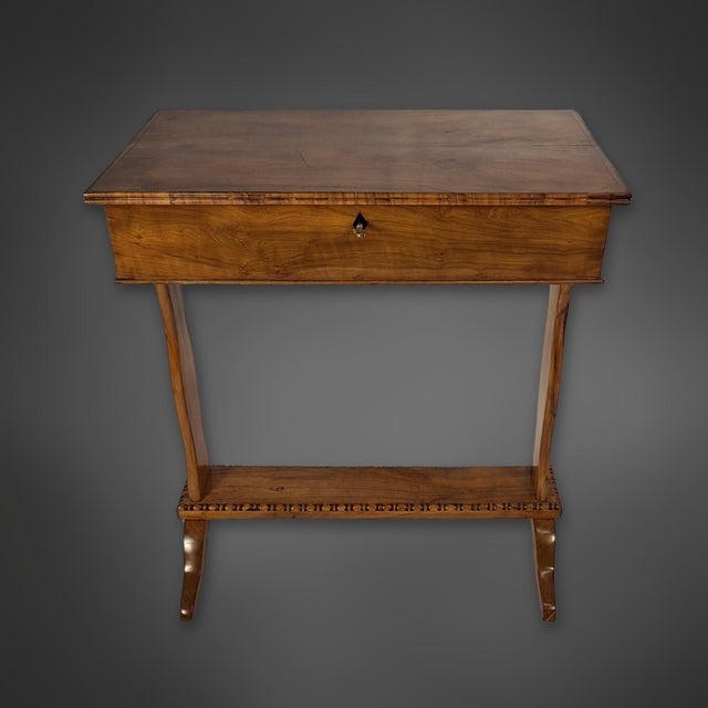 A Biedermeier-era sewing table made of solid cherry wood. The rectangular cabinet rests on two curved sides, which stand...