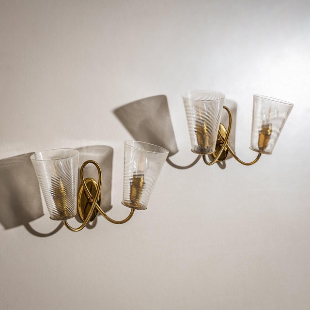 Pair of wall lights with loop-shaped brass mounts and conical clear glass shades featuring diagonal ribbed decoration....