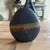 A sculptural ceramic bud vase featuring a flattened organic silhouette finished in a matte charcoal glaze with a flowing...