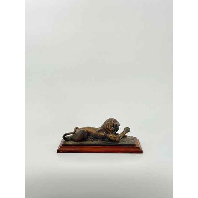 This striking bronze sculpture depicts a lion at rest, lying down while gently raising one powerful paw—a subtle yet...