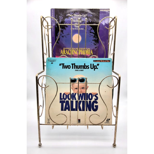 This vintage Mid-Century Modern wire magazine rack features a gold-tone finish, balled feet, and ornate scrollwork....