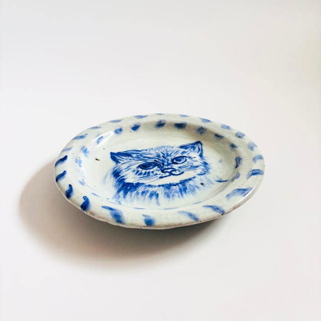 A vintage studio pottery cat plate. Features a blue hand painted cat face in the center with a pale gray base glaze.