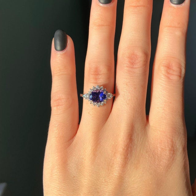 A tanzanite takes centre stage in a deep violet-blue at the heart of this solid 14K white gold ring, framed by sixteen...