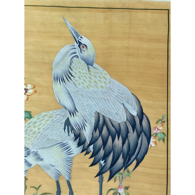 This painting depicts two elegant cranes standing amid flowering plants against a warm, golden background. The taller...