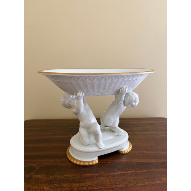 A neoclassical porcelain compote featuring two sculptural putti supporting a fluted bowl, finished with restrained gilt...