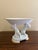 A neoclassical porcelain compote featuring two sculptural putti supporting a fluted bowl, finished with restrained gilt...