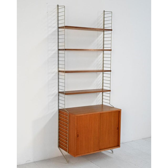 "String" bookcase in teak and lacquered metal with a suspended cabinet with sliding doors and adjustable shelves. String...