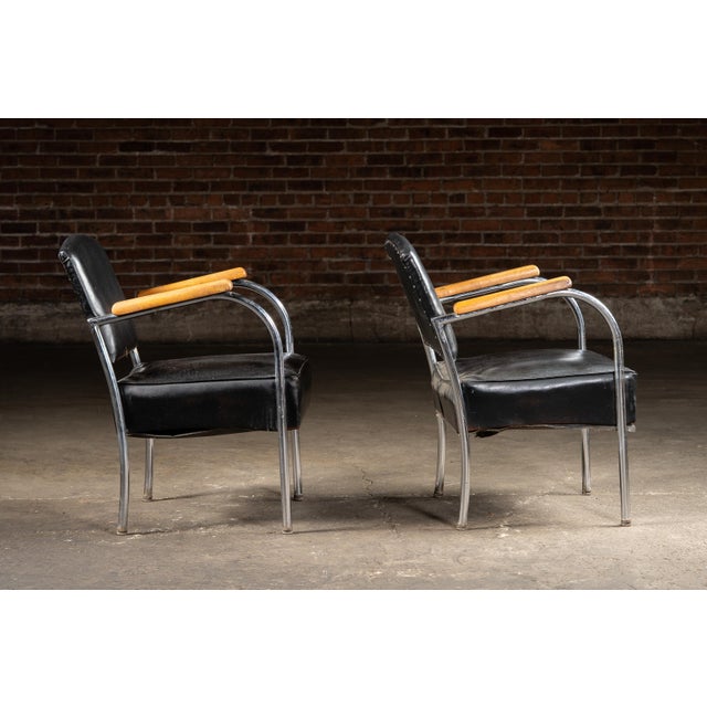Matched pair of Art Deco armchairs featuring tubular chrome frames, bent plywood armrests, and black vinyl upholstery. A...