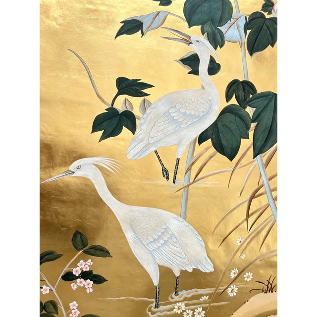An elegant hand painted gold leaf wallpaper featuring graceful white cranes surrounded by delicate flowering branches and...