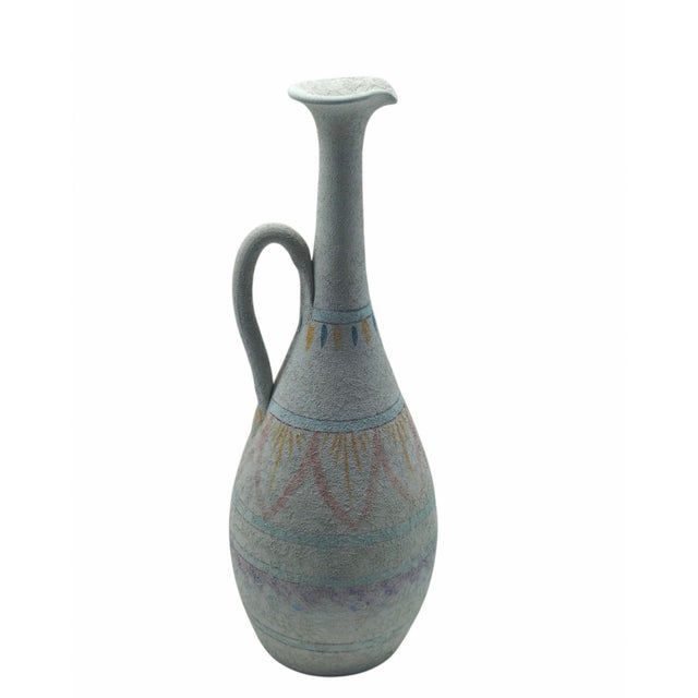 A pitcher-shaped vase with a handle, crafted from ceramic with a distinctive rough, textured finish that has a chalky...