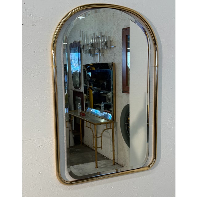 This is a vintage-style wall mirror featuring a polished brass frame with a rounded top and curved edges. The frame has a...