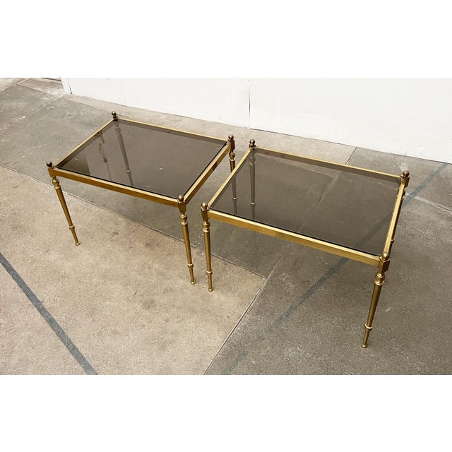 Two mid-century side tables in a neoclassical style. The tables consist of a brass frame with a table surface made of...
