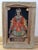 This piece displays deep black fabric surrounding a figure rendered in warm red, cream, and muted green threads, with the...