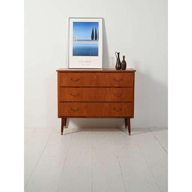 1950s-1960s teak chest of drawers with mahogany inserts, slender legs with brass tips, and metal handles with an Art Deco...