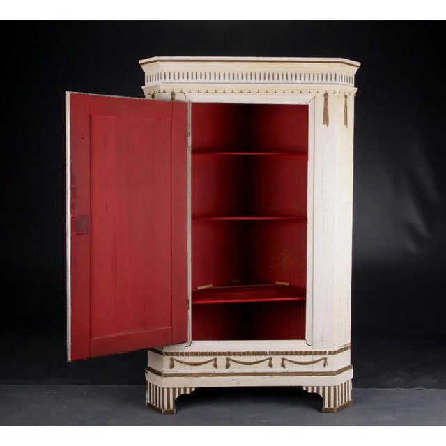 This Swedish corner cabinet, produced in the early 1800s, contains a series of gold-colored decorative features typical of...