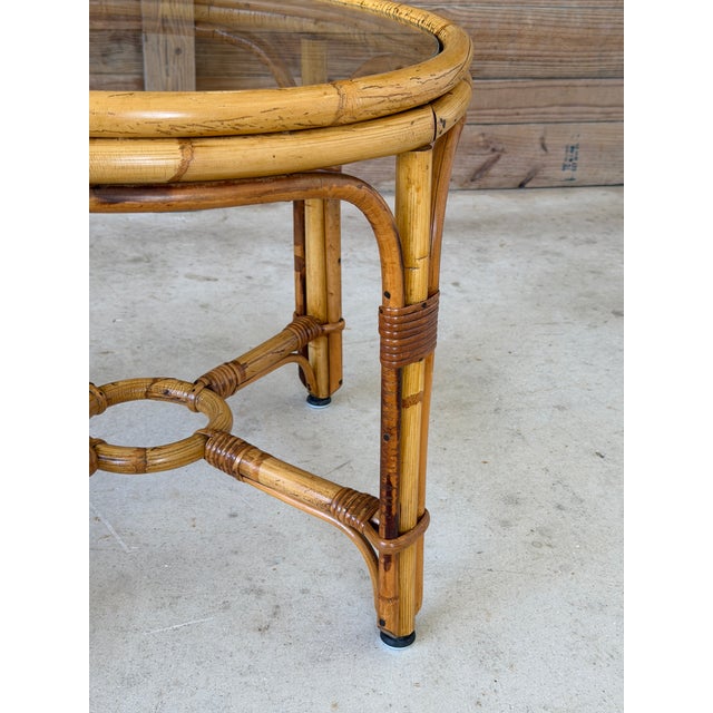 A charming vintage rattan side table featuring a classic round form, sculptural arched supports, and decorative wrapped...