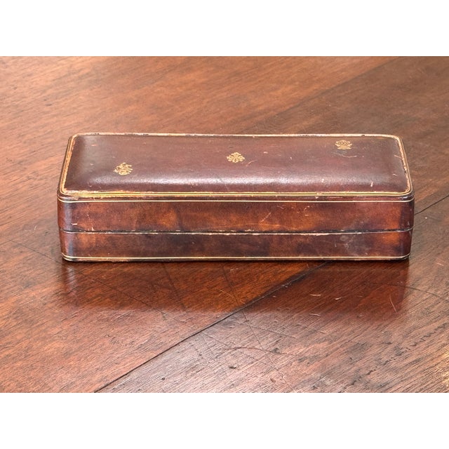 A rectangular brown leather box featuring gilt-tooled crests on the lid and a fine gold border line. The interior is lined...